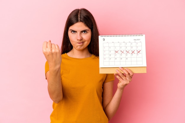 ピンクの背景にカレンダーを保持している若い白人女性が、カメラに拳を見せ、積極的な表情を見せている。