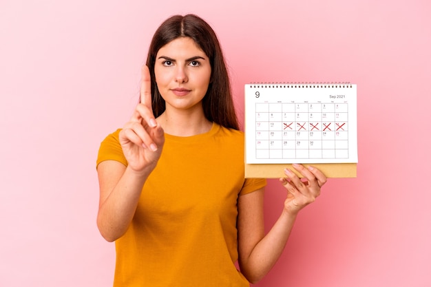 Foto giovane donna caucasica che tiene il calendario isolato su sfondo rosa che mostra il numero uno con il dito.