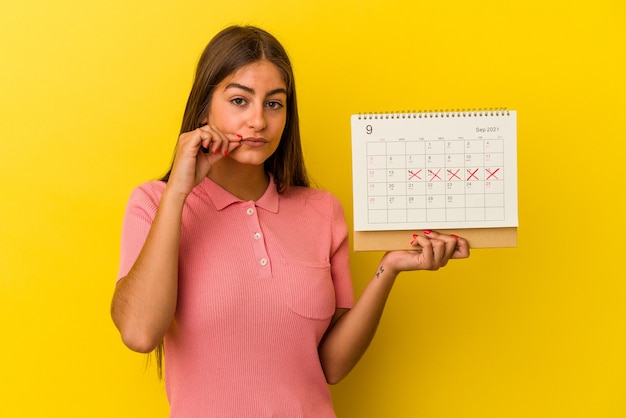 Young caucasian woman holding a calendar isolated on yellow wall with fingers on lips keeping a secret.