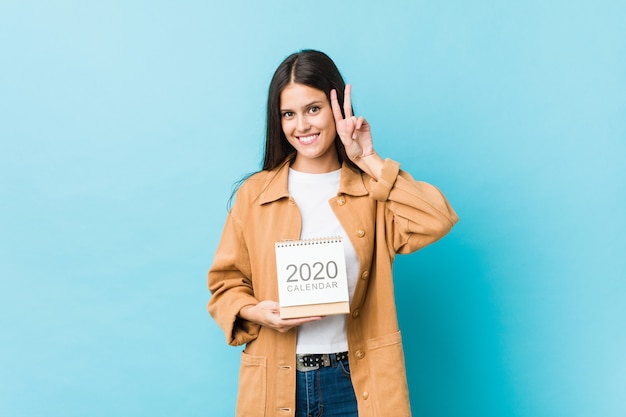 Young caucasian woman holding a calendar showing victory sign and smiling broadly.