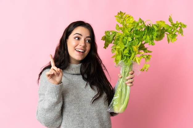 Young caucasian woman holding a celery isolated on blue wall intending to realizes the solution while lifting a finger up