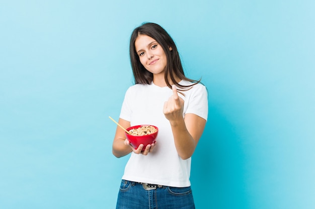 Young caucasian woman holding a cereals bowl pointing with finger at you as if inviting come closer