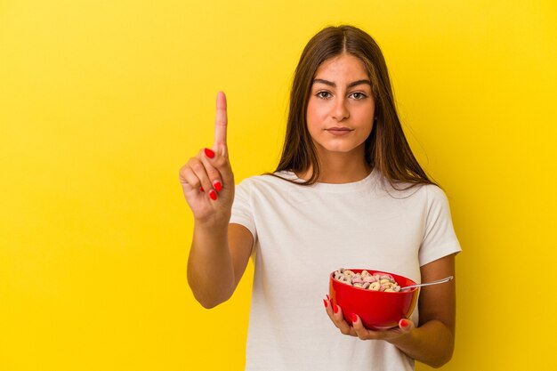 指でナンバーワンを示す黄色の背景に分離された穀物を保持している若い白人女性。