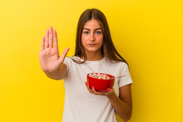 Young caucasian woman holding cereals isolated on yellow background standing with outstretched hand showing stop sign, preventing you.