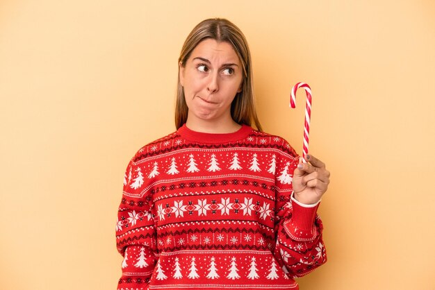 Foto la giovane donna caucasica che tiene un bastone di natale isolato su sfondo giallo confuso, si sente dubbiosa e insicura.