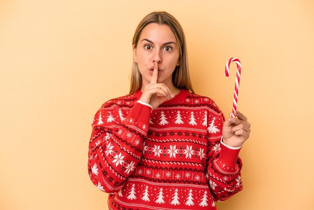 Foto giovane donna caucasica che tiene un bastone di natale isolato su sfondo giallo mantenendo un segreto o chiedendo silenzio.