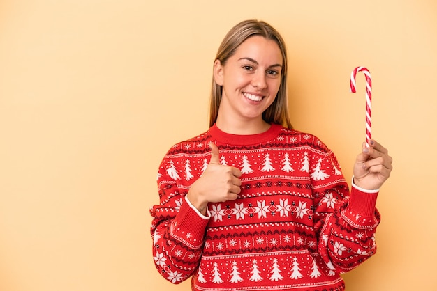 Young caucasian woman holding a christmas stick isolated on yellow background smiling and raising thumb up