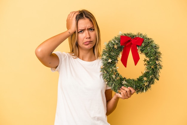 ショックを受けている黄色の背景に分離されたクリスマスリースを保持している若い白人女性、彼女は重要な会議を思い出しました。