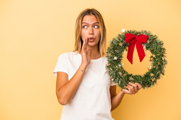 Young caucasian woman holding a christmas wreath isolated on yellow background is saying a secret hot braking news and looking aside