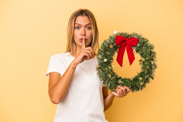 Young caucasian woman holding a christmas wreath isolated on yellow background keeping a secret or asking for silence.