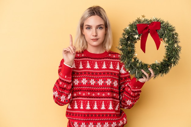 Young caucasian woman holding a Christmas wreath isolated on yellow background showing number one with finger.