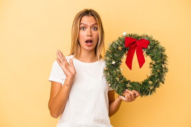 Young caucasian woman holding a christmas wreath isolated on yellow background surprised and shocked.