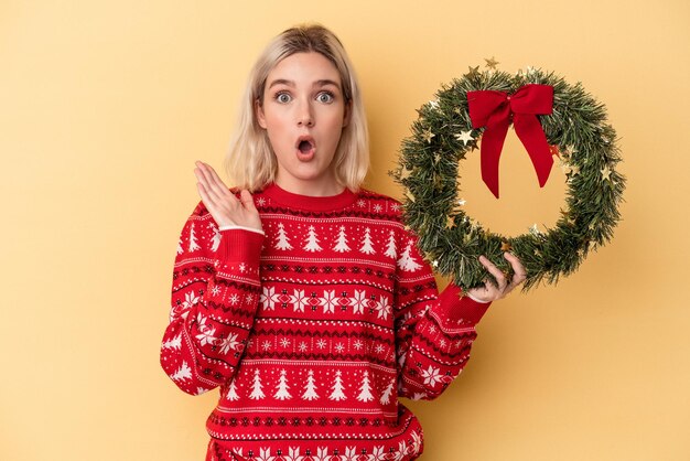 Young caucasian woman holding a Christmas wreath isolated on yellow background surprised and shocked.