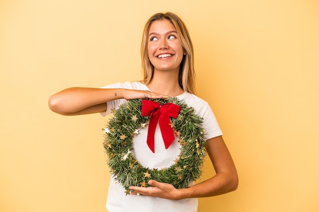 Young caucasian woman holding a christmas wreath isolated on yellow background