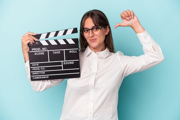 Young caucasian woman holding clapperboard isolated on blue background feels proud and self confident, example to follow.