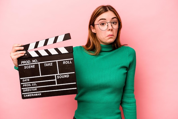 Young caucasian woman holding a clapperboard isolated on pink background shrugs shoulders and open eyes confused