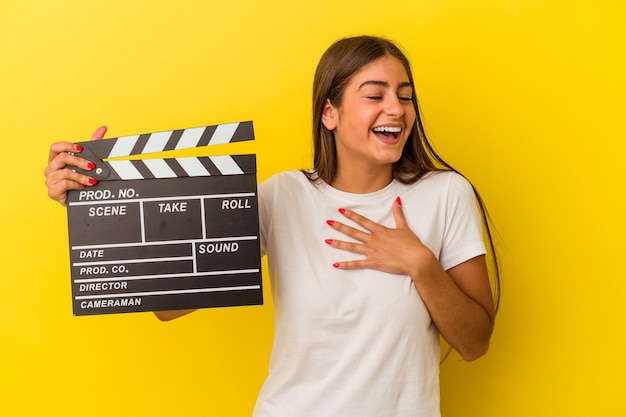 Young caucasian woman holding clapperboard isolated on white background laughs out loudly keeping hand on chest