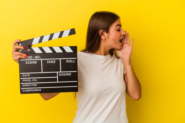 Young caucasian woman holding clapperboard isolated on white background shouting and holding palm near opened mouth.