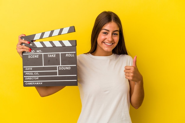 Young caucasian woman holding clapperboard isolated on white background smiling and raising thumb up