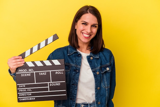 Young caucasian woman holding a clapperboard isolated on yellow background happy smiling and cheerful