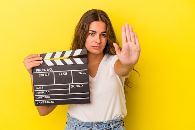 Young caucasian woman holding clapperboard isolated on yellow background  standing with outstretched hand showing stop sign, preventing you.