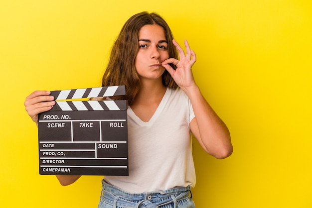 Young caucasian woman holding clapperboard isolated on yellow background  with fingers on lips keeping a secret.