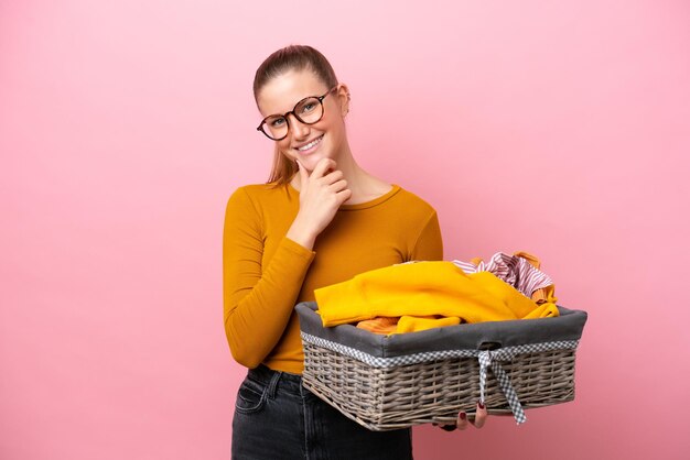 Young caucasian woman holding a clothes basket isolated on pink background happy and smiling