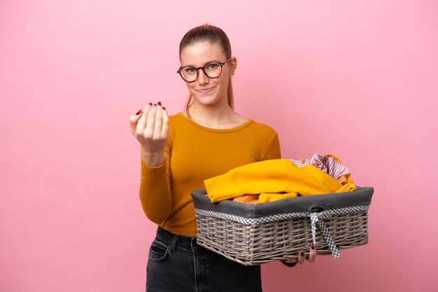 ピンクの背景に分離された服のバスケットを保持している若い白人女性手で来るように誘う