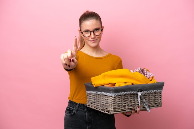 ピンクの背景で隔離の服のバスケットを保持し、指を見せて持ち上げる若い白人女性