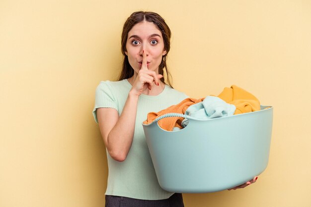 Young caucasian woman holding a clothes basket isolated on yellow background keeping a secret or asking for silence