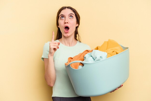 Young caucasian woman holding a clothes basket isolated on yellow background pointing upside with opened mouth