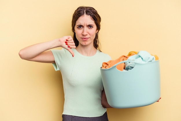 嫌いなジェスチャーを示す黄色の背景に分離された服のバスケットを持っている若い白人女性、親指を下に向けます。不一致の概念。