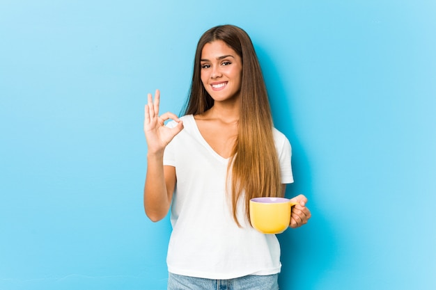 Молодая кавказская женщина держа кружку кофе жизнерадостную и уверенно показывая одобренный жест.