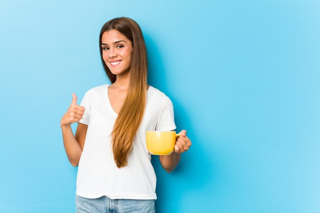 Giovane donna caucasica che tiene una tazza di caffè che sorride e che alza il pollice in su