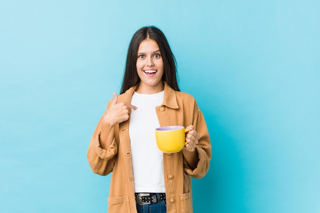 Young caucasian woman holding a coffee mug surprised pointing at himself, smiling broadly.