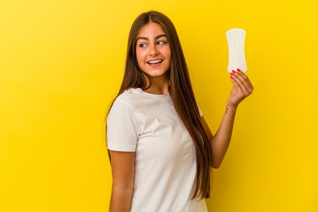 Young caucasian woman holding a compress isolated on yellow background looks aside smiling, cheerful and pleasant