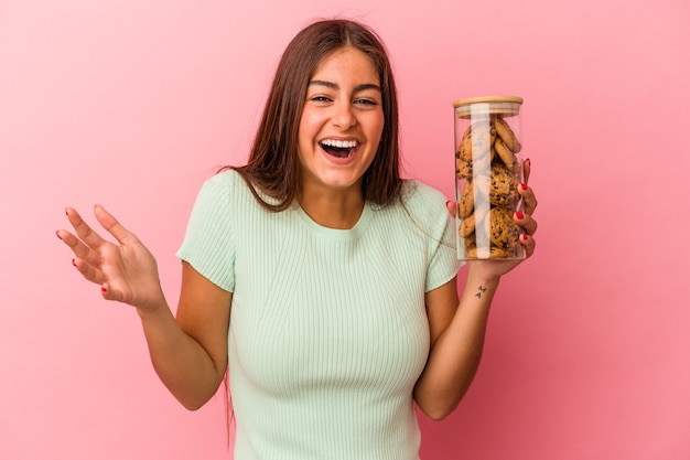 Young caucasian woman holding a cookies jar isolated on pink background receiving a pleasant surprise, excited and raising hands.