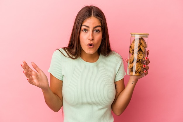 Photo young caucasian woman holding a cookies jar isolated on pink background surprised and shocked.