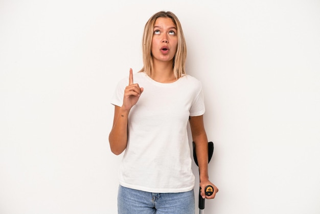 Young caucasian woman holding crutch isolated on white background pointing upside with opened mouth.