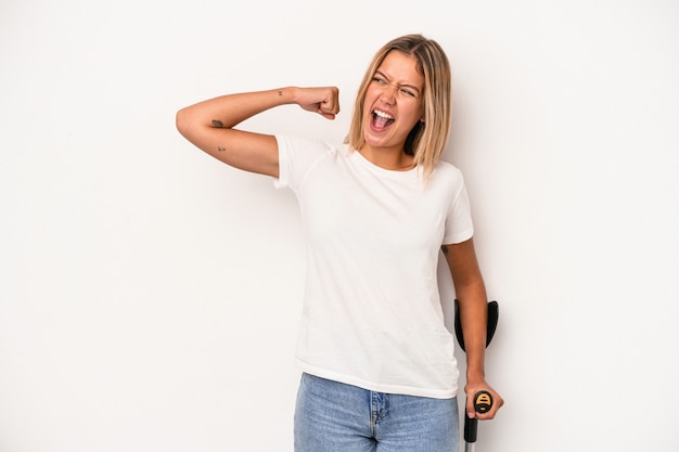 Foto giovane donna caucasica che tiene stampella isolata su sfondo bianco alzando il pugno dopo una vittoria, concetto vincitore.