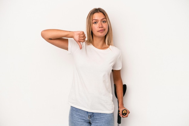 Photo young caucasian woman holding crutch isolated on white background showing a dislike gesture, thumbs down. disagreement concept.