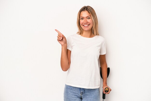 Young caucasian woman holding crutch isolated on white background smiling and pointing aside, showing something at blank space.