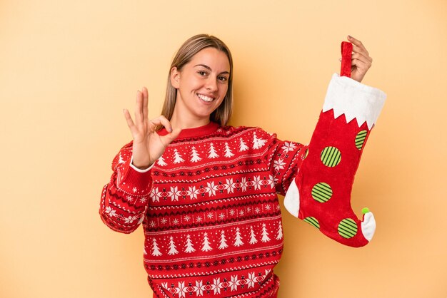 Young caucasian woman holding a elf sock isolated on yellow background cheerful and confident showing ok gesture.