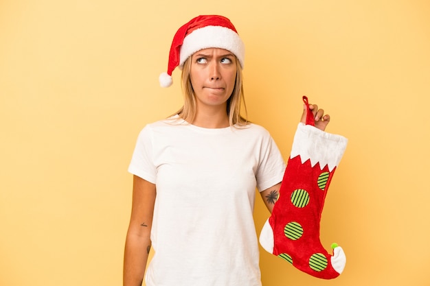 Young caucasian woman holding a elf sock isolated on yellow background confused, feels doubtful and unsure.