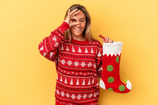Photo young caucasian woman holding a elf sock isolated on yellow background excited keeping ok gesture on eye.