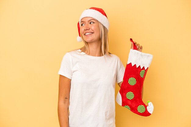 Young caucasian woman holding a elf sock isolated on yellow background looks aside smiling, cheerful and pleasant.