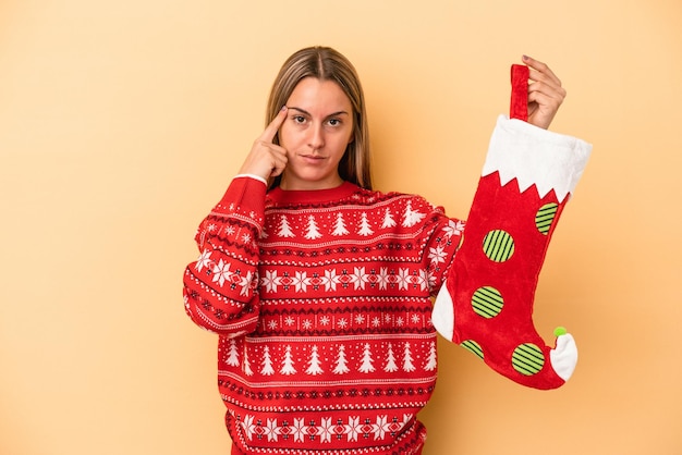 Photo young caucasian woman holding a elf sock isolated on yellow background pointing temple with finger, thinking, focused on a task.