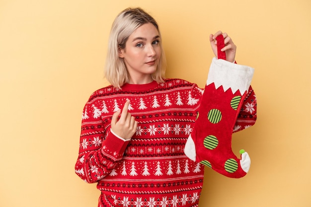 Young caucasian woman holding a elf sock isolated on yellow background pointing with finger at you as if inviting come closer.