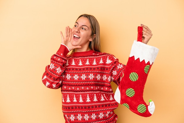 Young caucasian woman holding a elf sock isolated on yellow background shouting and holding palm near opened mouth.