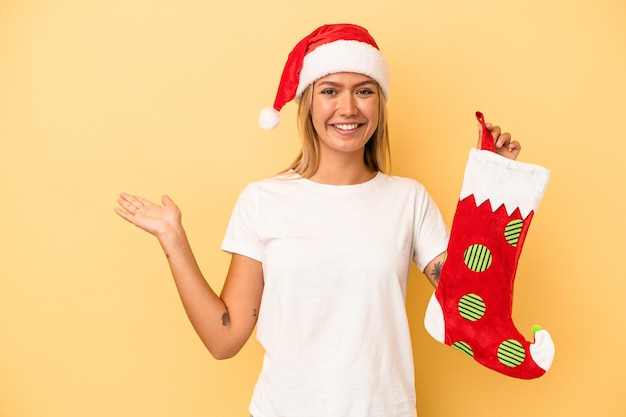 Young caucasian woman holding a elf sock isolated on yellow background showing a copy space on a palm and holding another hand on waist.